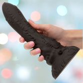 A hand with pink-painted nails holds the CalExotics Monster Dong Frankenstein’s Monster, a large textured black silicone fantasy-themed adult toy, against a blurred background with colorful bokeh lights.